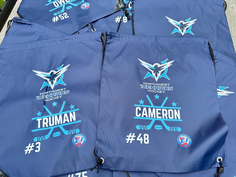 May include: A collection of navy blue drawstring bags featuring the Northwest Icehawks Hockey logo in white and light blue. Each bag is personalized with a player's name, such as Truman or Cameron, and a jersey number like #3 or #48.