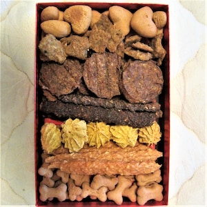 May include: A red box filled with a variety of dog treats. The treats are arranged in layers and include biscuits, chews, and bones.