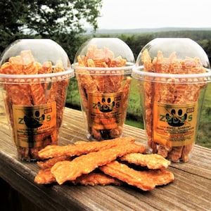 Op de afbeelding: Drie doorzichtige plastic bekers gevuld met oranje hondenkoekjes. De bekers hebben een gouden label met de tekst "Zeus's Gourmet Bakery" en een poot afdruk logo. De koekjes liggen op een houten oppervlak.