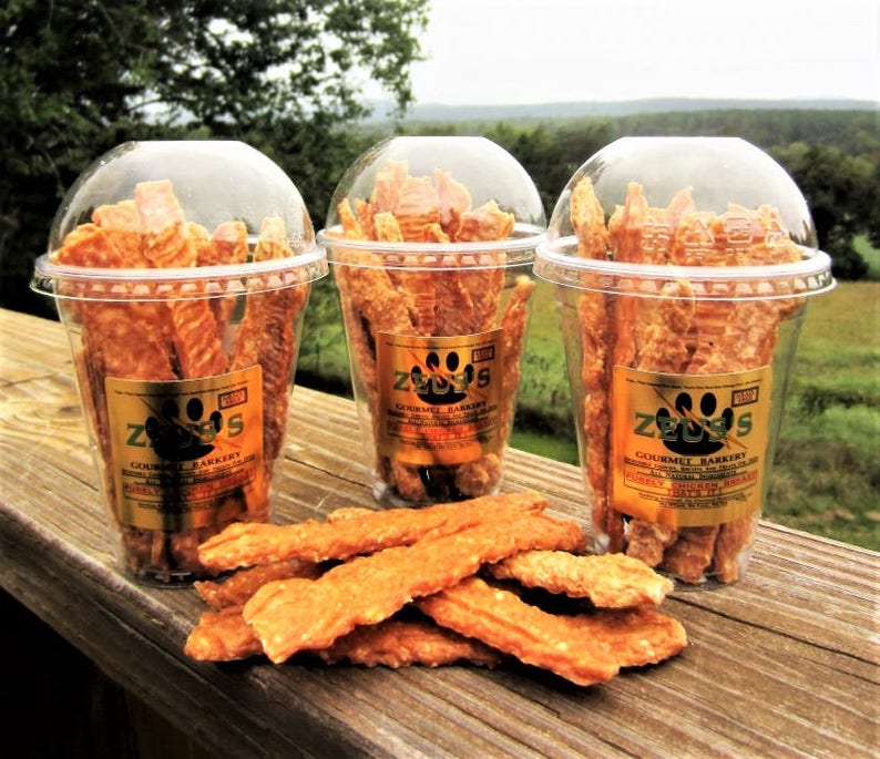 Op de afbeelding: Drie doorzichtige plastic bekers gevuld met oranje hondenkoekjes. De bekers hebben een gouden label met de tekst "Zeus's Gourmet Bakery" en een poot afdruk logo. De koekjes liggen op een houten oppervlak.
