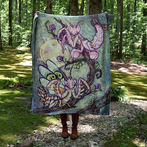 May include: A soft, plush throw blanket featuring a whimsical illustration of two praying mantises on a vine with pink flowers and a full moon in the background. The mantises are depicted in a cartoon style with large eyes and detailed wings.