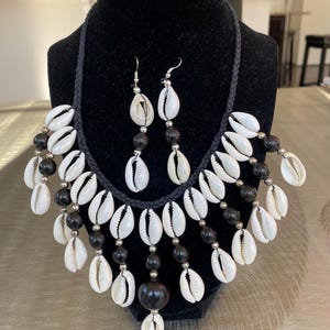 May include: A black necklace and earring set displayed on a black mannequin. The necklace features white seashells, black beads, and silver-colored accents. The earrings have matching seashells and black beads.