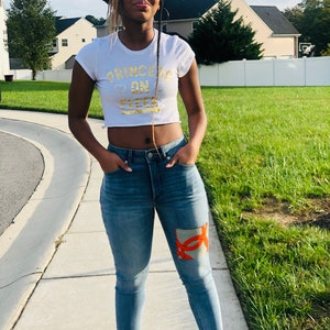 May include: A white crop top with the text "PRINCESS ON FLEEK" in gold lettering. The model is wearing light blue jeans with an orange and beige patch on the right knee. The model is standing on a sidewalk.