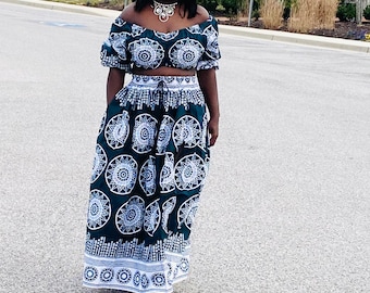May include: A woman wearing a green and white off-the-shoulder crop top and matching maxi skirt with a white and black geometric pattern. The skirt has a white waistband and a white trim around the bottom.