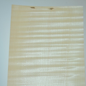 May include: A sheet of light wood veneer with a wavy grain pattern. The veneer is light beige in colour and has a smooth, polished surface.