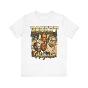 May include: White t-shirt with a vintage-style graphic of Robert Mugabe, a former president of Zimbabwe. The graphic features a collage of images of Mugabe and other figures, with the text "ROBERT MUGABE" in gold lettering.