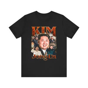 May include: Black t-shirt featuring a vintage-style graphic of Kim Jong Un. The graphic includes the name "KIM" in large letters and "Jong Un" below, along with multiple images of Kim Jong Un.