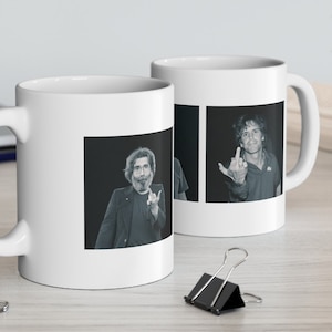 May include: Two white ceramic mugs with black and white images of a man. One mug features a man with a beard and long hair, making a hand gesture. The other mug features a man with short hair, making a hand gesture.