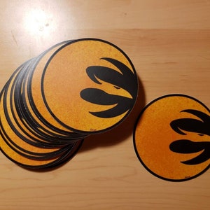 Könnte beinhalten: Ein Stapel runder orangefarbener und schwarzer Aufkleber mit einem stilisierten Vogeldesign. Die Aufkleber befinden sich auf einer Holzoberfläche.