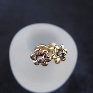 May include: A pair of gold-coloured flower stud earrings. Each earring features a six-petal flower design with a small, dark-coloured stone in the centre. The earrings are displayed on a white, rounded surface against a dark background.