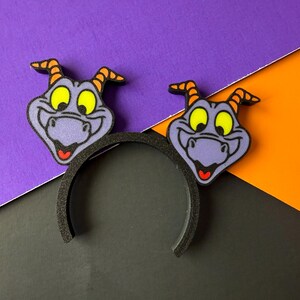 May include: A black headband with two purple, yellow, orange, and red cartoon character cutouts. The background is a diagonal split of purple, orange, and black.