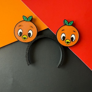 May include: A black headband with two orange cartoon orange character decorations. The orange characters have green leaves, white eyes, and yellow beaks. The background is a diagonal split of orange, red, and black.