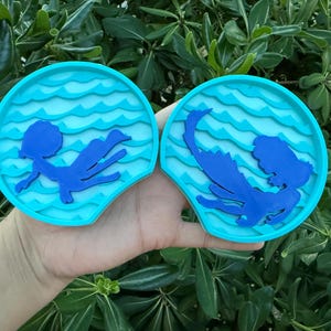 May include: Two blue and turquoise mermaid-themed ear headbands. Each headband features a mermaid silhouette swimming in a wavy ocean design. The headbands are held in a hand against a background of green leaves.