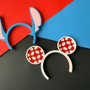 May include: Two headbands, one blue with pink ears and the other white with red and white circular ears. The headbands are on a background of red, blue, and black geometric shapes.