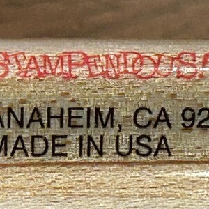 May include: Close-up of a wooden stamp with the text "FUN STAMPS STAMPENDOUS" in a black and white logo. The text "STAMPENDOUS" is in red. Below is "ANAHEIM, CA 92805 MADE IN USA" in black text. The copyright symbol and the year 1996 are also visible.