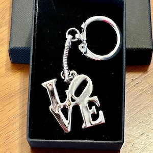 May include: A silver keychain with a stylized "LOVE" charm. The keychain is in a black velvet box.