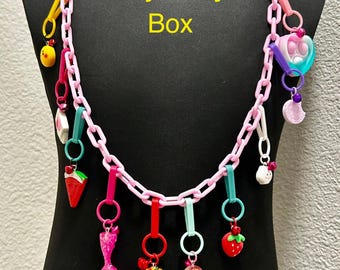 May include: A pink plastic chain necklace with various colorful charms, including a mermaid tail, a strawberry, a hamburger, a rainbow, a watermelon slice, a heart, a chick, a cloud, and a candy. The necklace is displayed on a black mannequin and the text "Mystery Box" is written in yellow on the mannequin's chest.