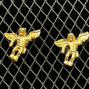 Pode incluir: Um par de brincos de anjo de ouro. Cada brinco apresenta um pequeno anjo com as asas abertas, segurando uma pequena esfera em uma m&atilde;o.