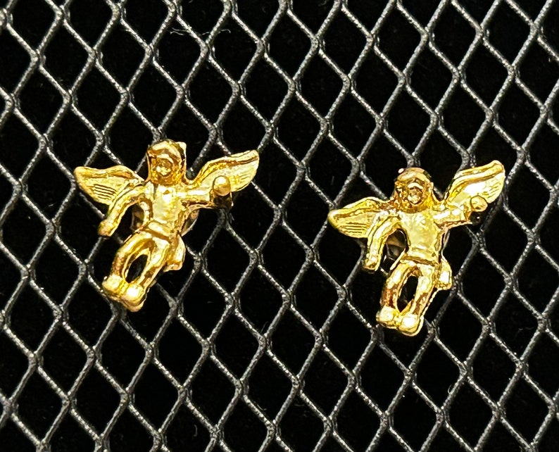 Pode incluir: Um par de brincos de anjo de ouro. Cada brinco apresenta um pequeno anjo com as asas abertas, segurando uma pequena esfera em uma m&atilde;o.