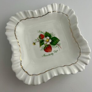 May include: A square white porcelain dish with a ruffled edge and a thin gold trim. The center features a botanical illustration of red strawberries, green leaves, and a white flower, with the green cursive text Strawberry Ripe below it.
