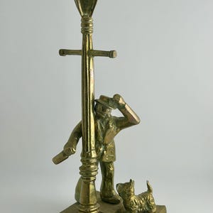 May include: A polished brass figurine depicting a person in a coat and hat standing next to a tall street lamp, holding a small object in one hand and tipping their hat with the other, accompanied by a small dog standing on a rectangular base.