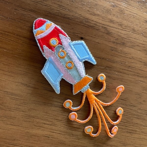 Könnte beinhalten: Ein bunter gestickter Aufnäher mit einer Rakete mit orangefarbenen Tentakeln. Die Rakete ist rot, weiß und blau mit orangefarbenen Akzenten. Die Tentakel sind orange mit weißen und rosa Akzenten.