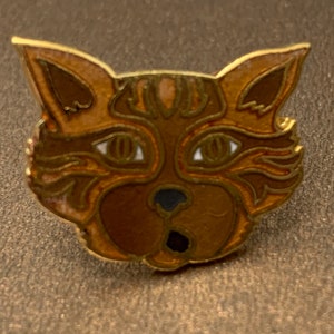 May include: A gold-colored metal pin with a detailed design of a cat's face. The cat has brown fur with white accents around the eyes and nose.
