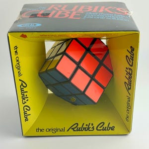 Puede incluir: El rompecabezas original del Cubo de Rubik en su caja original. El cubo tiene lados rojos, naranjas, azules, amarillos, verdes y blancos. La caja es amarilla con texto negro y un logotipo azul Ideal.