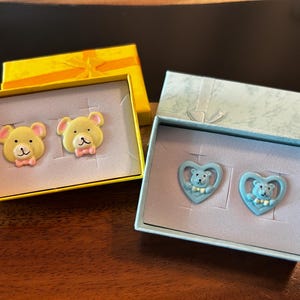 Könnte beinhalten: Zwei Paar Ohrringe in Geschenkboxen. Ein Paar zeigt gelbe Bärengesichter mit rosa Schleifen, das andere Paar hat herzförmige blaue Ohrringe mit Bärenmotiven. Die Schachteln sind gelb und hellblau.
