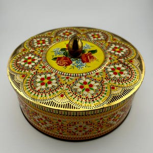 May include: A round, vintage tin box with a decorative floral design. The lid features a yellow centre with roses and blue flowers, topped with a gold knob. The box has a gold rim and a detailed, colourful pattern.