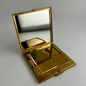May include: A gold-toned compact mirror, open to reveal a mirror and a compartment. The compact has a rectangular shape with rounded corners and a small clasp. The mirror reflects the surrounding environment.