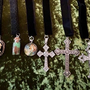 May include: Six silver pendants on black ribbon necklaces. The pendants include a cameo with a woman's face, a small glass bottle with green and brown contents, a round pendant with a colorful image, and three silver crosses of different designs.