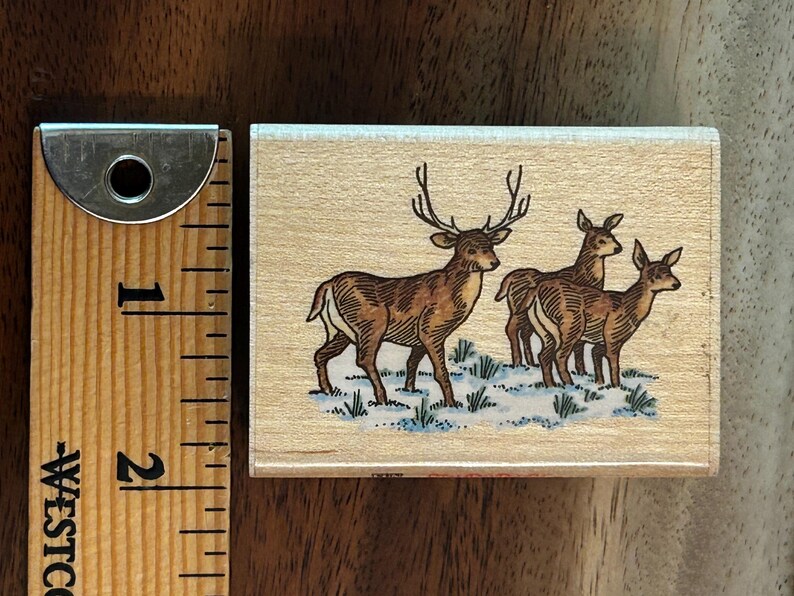 May include: A rectangular wooden stamp featuring a brown illustration of three deer in a snowy field. The stamp measures approximately 2 inches in length and is next to a ruler.