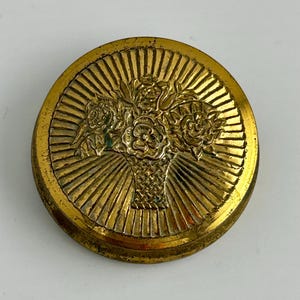 May include: A round, gold-colored button with a raised floral design. The button features a basket of roses in the center, surrounded by a sunburst pattern. The button is likely made of metal and has a vintage aesthetic.