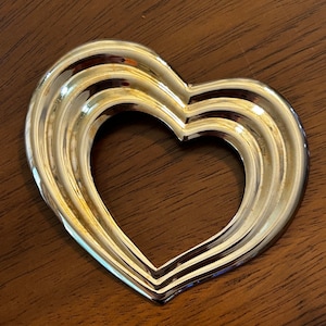 May include: A shiny, silver-colored heart-shaped object with a layered design. The heart has a hollow center and is set against a dark brown wooden surface. The object reflects light, creating highlights and shadows.