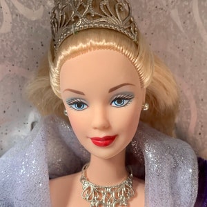 Peut inclure: Une poupée en porcelaine aux cheveux blonds portant une couronne et un collier en argent. Elle porte une robe violette et argentée avec un châle blanc.