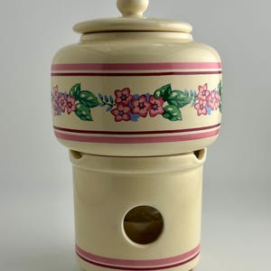 May include: A cream-colored ceramic butter keeper with a lid. The upper section features a horizontal band of pink flowers and green leaves between two thin pink stripes. The base has a circular cutout and matching pink stripes at the bottom.