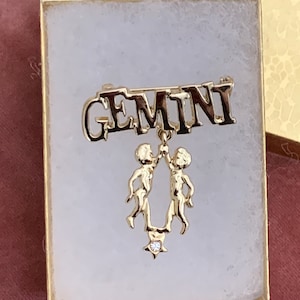 May include: Gold-tone Gemini zodiac sign brooch with a dangling charm of two figures holding hands and a rhinestone star.