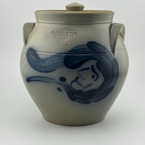 Op de afbeelding: Een grijze steengoed pot met een deksel, versierd met blauwe patronen. De pot heeft een ronde vorm, twee handvatten en een deksel met een kleine knop. De tekst "Westbrook Pottery Columbus, NY 1991" is zichtbaar aan de zijkant.
