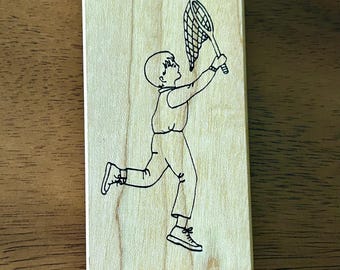 May include: A wooden stamp featuring a black line drawing of a boy running with a net in his hand. The boy is looking up at the net.
