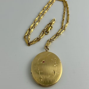 May include: A gold-tone oval locket pendant suspended from a matching textured link chain. The locket features a brushed gold finish with engraved swirling vine patterns and a single small red stone accent embedded in the center of the design.