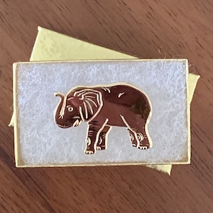 May include: A small, white box with a gold trim and a brown elephant design. The elephant is facing right and has a trunk raised in the air.