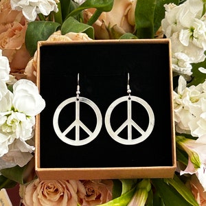 Op de afbeelding: Een paar zilveren peace-teken oorbellen in een zwarte geschenkdoos. De oorbellen zijn op een witte achtergrond met bloemen.