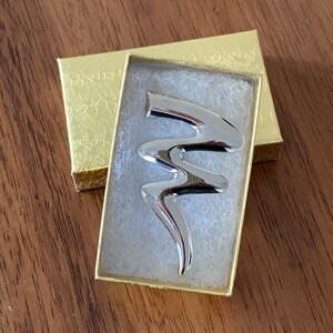 May include: A silver abstract design brooch in a small white and gold box.