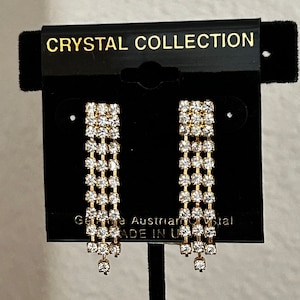 May include: A pair of gold tone dangle earrings with clear crystal stones. The earrings are on a black card with the text "CRYSTAL COLLECTION" and "Genuine Austrian Crystal MADE IN U.S.A."