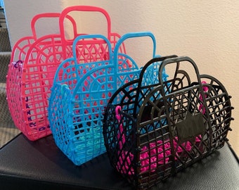 May include: Four plastic tote bags in different colors. The bags are made of a woven plastic material and have a handle on top. The bags are pink, blue, black, and clear.