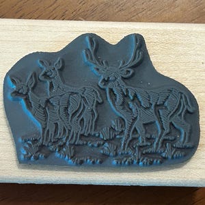 May include: A wooden stamp featuring a detailed illustration of three deer in a grassy field. The stamp's design is in dark blue, mounted on a rectangular wooden block. The deer have intricate antlers and body details.