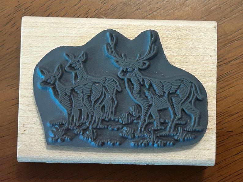 May include: A wooden stamp featuring a detailed illustration of three deer in a grassy field. The stamp's design is in dark blue, mounted on a rectangular wooden block. The deer have intricate antlers and body details.