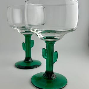 May include: Two clear margarita glasses with green cactus-shaped stems and bases. The stems have a ribbed texture and the base is a solid green color. The glasses are made of clear glass.