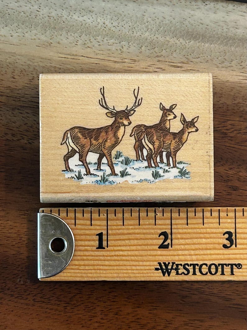 May include: A rectangular wooden stamp featuring a brown deer with large antlers standing in a snowy field, with three other deer behind it. The stamp is approximately 2.5 inches long, with a ruler in the foreground.